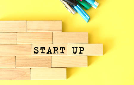 Wooden blocks stacked next to pens and pencils on a yellow background. STARTUP text on a wooden...
