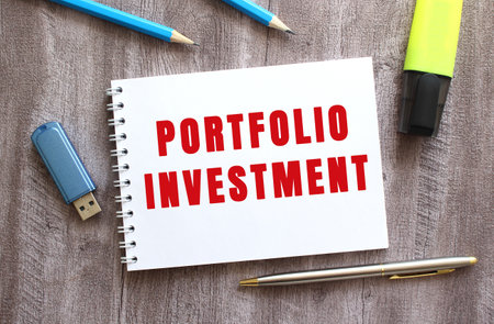 Top view of the workspace. Notepad with PORTFOLIO INVESTMENT text, pen, pencils, flash drive on gray wooden table. Business concept.の写真素材