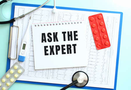 A notepad with the text ASK THE EXPERT lies on a blue medical clipboard with a stethoscope and pills on a blue background. Medical concept.の写真素材