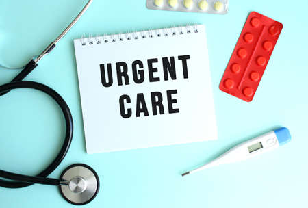The text URGENT CARE is written on a white notepad that lies next to the stethoscope and pills on a blue background. Medical conceptの写真素材