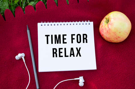 A notebook with the inscription TIME FOR RELAX. Nearby are headphones and an apple. Red plaid and green grass in the background. Concept for design.の写真素材