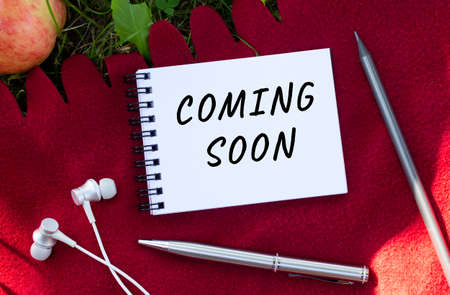 A notebook with the inscription COMING SOON. Nearby are headphones and an apple. Red plaid and green grass in the background. Concept for design.の写真素材
