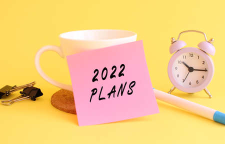 Pink paper with the text 2022 PLANS on a white cup. Clock, pen on a yellow background. Design concept.の写真素材