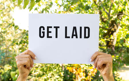 Female hands hold a sheet of white paper overhead. On the sheet the inscription GET LAID. Against the backdrop of nature. Information poster.の写真素材