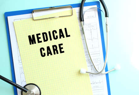 Blue medical clipboard with a sheet of yellow paper with the text MEDICAL CARE and a stethoscope.の写真素材