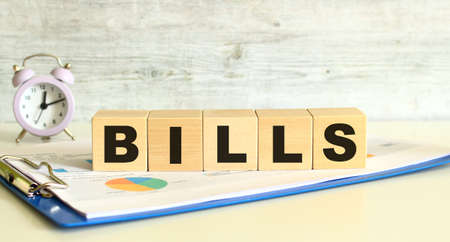 Wooden cubes lie on a folder with financial charts on a gray background. The cubes make up the word BILLS.の写真素材