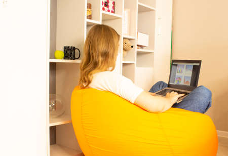 A young woman sits on an orange beanbag chair in the room and works using a laptop at home. Remote work concept, freelancing.の写真素材