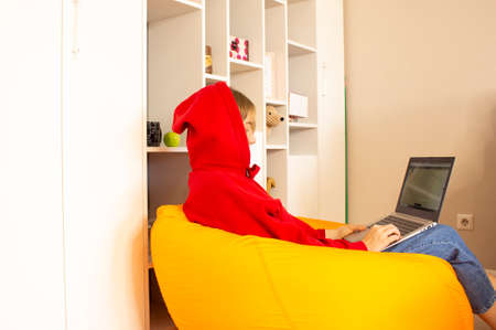 A young woman sits on an orange beanbag chair in the room and works using a laptop at home. Remote work concept, freelancing.の写真素材