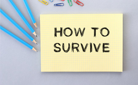 HOW TO SURVIVE text in notebook on gray background next to pencils and paper clips. concept.の写真素材