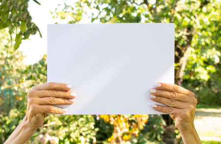 Female hands hold an empty sheet of white paper on the background of nature. information poster.の写真素材
