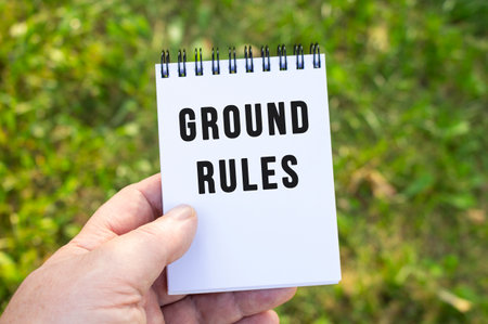 A man's hand holds a notebook with the inscription GROUND RULES. Man's hand with notepad on the background of nature. Close-up of an open notebook in the park.の写真素材