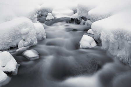 Winter landscape of mountain stream bursting out from under stones covered with caps of white snowの写真素材