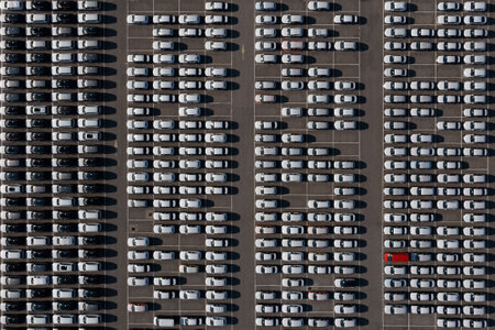 Top-down aerial view of a car storage area containing over 300 vehicles all black and white except one red standing in straight rows and columns looking like an abstract patternの写真素材
