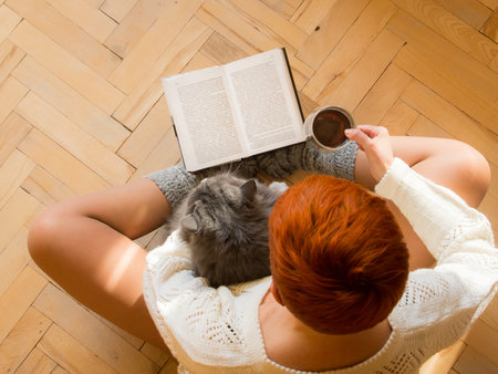 The red-headed girl is sitting on the wooden floor reading a book with a cup of tea in her right hand and a big furry cat in her left handの写真素材