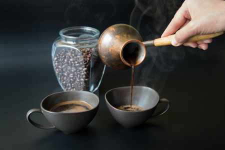 A hand pours black coffee from a vintage Turkish coffee pot into one of a pair of dark cups against a black backgroundの写真素材