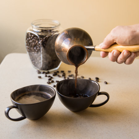 A hand pours black coffee from a vintage Turkish coffee pot into one of a pair of dark cups with a glass jar full of coffee beans out of focus in the background and a few grains scattered on the tableの写真素材