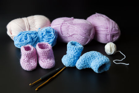 Crochet pink and blue booties for a newborn with a pair of crotchets in the foreground and balls of yarn in the background out of focus against a black backdropの写真素材