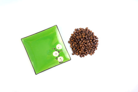 A pile of coffee beans next to a square green saucer, white backgroundの写真素材