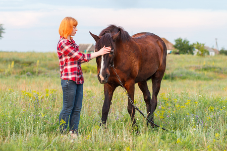 red-haired girl in a red plaid shirt stroking a brown horseの写真素材