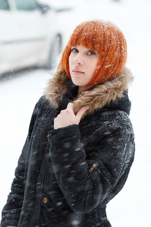 Winter portrait of a cute red-haired girl with blue eyes in a black jacket with fur during a snowstorm,  red-haired woman stands in the street during a heavy snowfall, look at the camera, verticalの写真素材