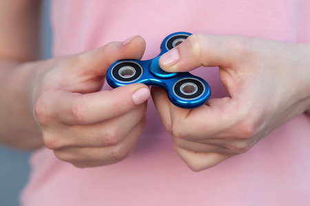 girl in pink t-shirt is playing blue metal spinner in hands on the street, woman playing with a popular fidget spinner toy, anxiety relief toy, anti stress and relaxation fidgetsの写真素材