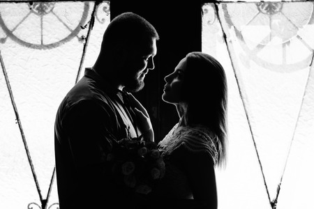 portrait of a romantic couple in a backlight from a window or door, silhouette of a couple in a doorway with a backlight, couple of lovers groom and bride at the window, black and white photography .の写真素材