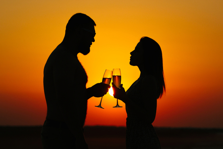 happy couple enjoying a glass of wine or champagne, silhouette of couple in love drinking wine from wineglasses during romantic dinner at sunset on the beach, sunset backgroundの写真素材