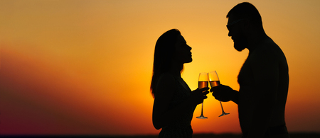 silhouettes of man and woman at sunset dramatic sky background, couple toasting wine glasses in romantic date setting, looking each other, smiling and holding in their hands glasses of champagneの写真素材