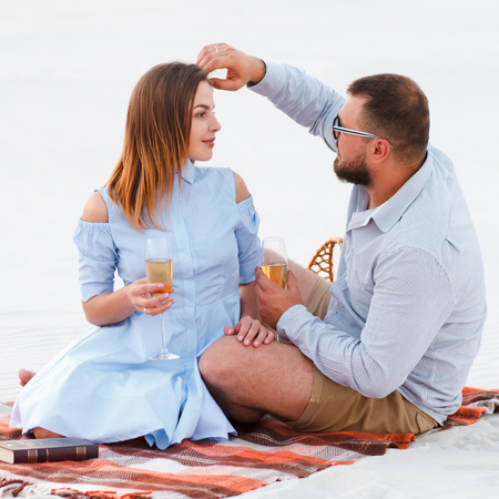 attractive couple sitting together, looking each other, happy couple enjoying picnic on the white sand beach and drinking wine or champagne during romantic dinner, happy couple sitting on a blanketの写真素材