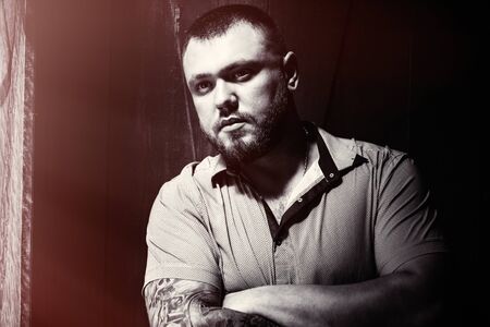 brutal bearded man with a tattoo on his arm, a portrait of a man in dramatic light against a brown wooden wall, attractive bearded male with tattoo on arm dressed in a shirt, man looks in the sideの写真素材