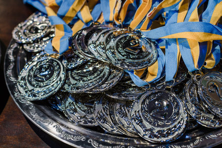 Many silver medals with blue yellow ribbons on a silver tray, awards of champions, sport achievements, second place, prize for the winnerの写真素材