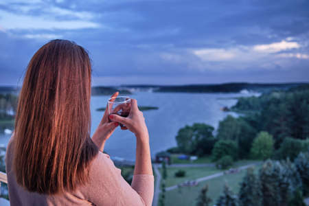 Back view of a happy free woman enjoying a beautiful serene morning, looking at the sea lake and landscape, starting a new day, drinking juice relaxing.の写真素材