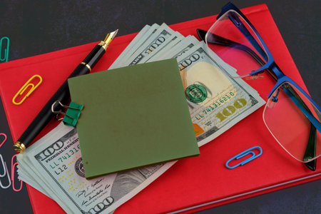 A sticker for writing attached to dollars on a red business notebook, glasses next to it, a pen on a...