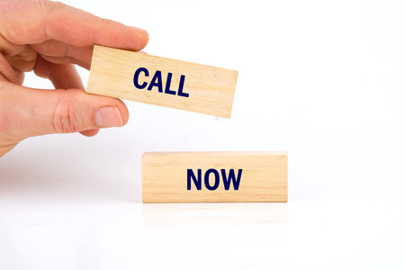 Conceptual call now symbol. Copy space. Call now message written laid out by a man's hand with wooden blocks on a white backgroundの写真素材