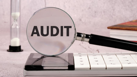 Copy space and business concept. A word AUDIT written through a magnifying glass on a gray background in a composition with a calculator, an hourglass and a bookの写真素材