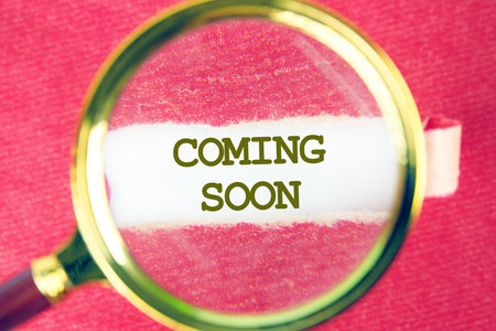 Conceptual coming soon symbol. Copy space. COMING SOON words written on white paper through a magnifying glass under a torn piece of red paperの写真素材