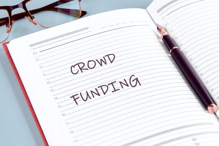 A notebook opened to a blank page features the term crowdfunding prominently written in bold...