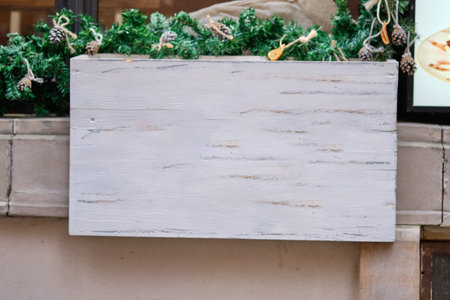 A blank wooden sign is positioned in front of lush greenery and festive decorations, enhancing the inviting atmosphere of a bustling urban restaurant during the holiday season.の写真素材