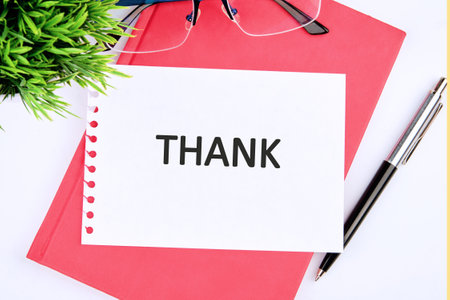 A thank you card is placed on top of a pink notebook, with a pair of glasses and a pen nearby. This setup creates a warm and inviting workspace atmosphere for gratitude notes.の写真素材