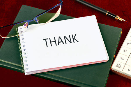 A thank you note lies open on a green notebook, accompanied by a pair of glasses and a pen, reflecting a moment of appreciation on a dark wooden surface.の写真素材