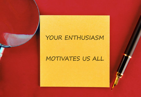 A bright yellow sticky note displays an uplifting message about enthusiasm. A pen and magnifying glass are placed beside it on a bright red surface.の写真素材