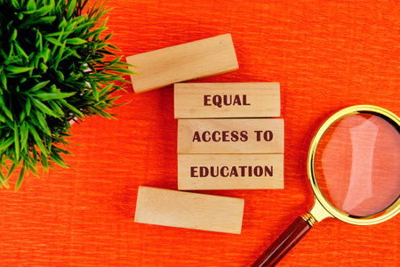 Wooden blocks spelling equal access to education sit on a bright orange surface beside a magnifying glass and a small plant, emphasizing the importance of inclusive education.の写真素材