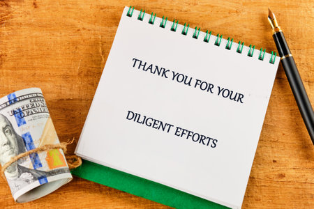 A note expressing gratitude for diligent efforts rests on a wooden desk near rolled currency and a pen, highlighting recognition of hard work and dedication.の写真素材