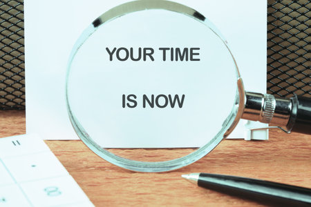 Motivational concept words YOUR TIME IS NOW focused on the notebook through a magnifying glass.の写真素材