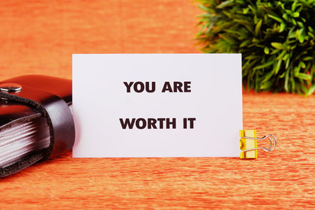 YOU ARE WORTH IT words written on a card on a bright orange table next to a black notebook and a small green plant.の写真素材