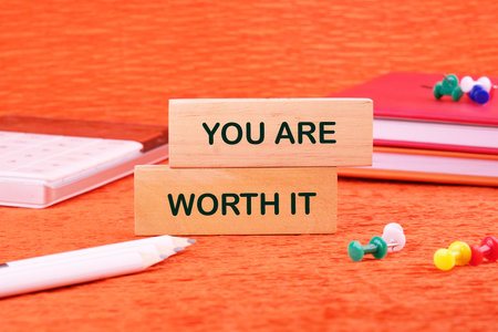 YOU ARE WORTH IT words made of wooden blocks on an orange background with stationery lying next to it.の写真素材