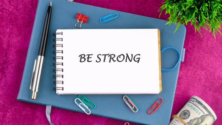 A notepad with the words BE STRONG prominently displayed is placed on a blue surface. Colorful...