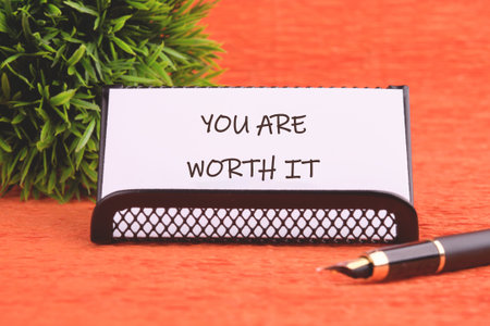 YOU ARE WORTH IT words written on the business card holder. Next to it is a fountain pen and a small ornamental plant on an orange background.の写真素材