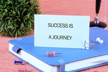 Concept text SUCCESS IS A JOURNEY written on a card on a blue notebook next to colorful pins and an ornamental plant.の写真素材