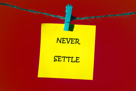 Concept words NEVER SETTLE written on a bright yellow note, which is attached to the shoelace with a blue clothespin on a bright red background.の写真素材